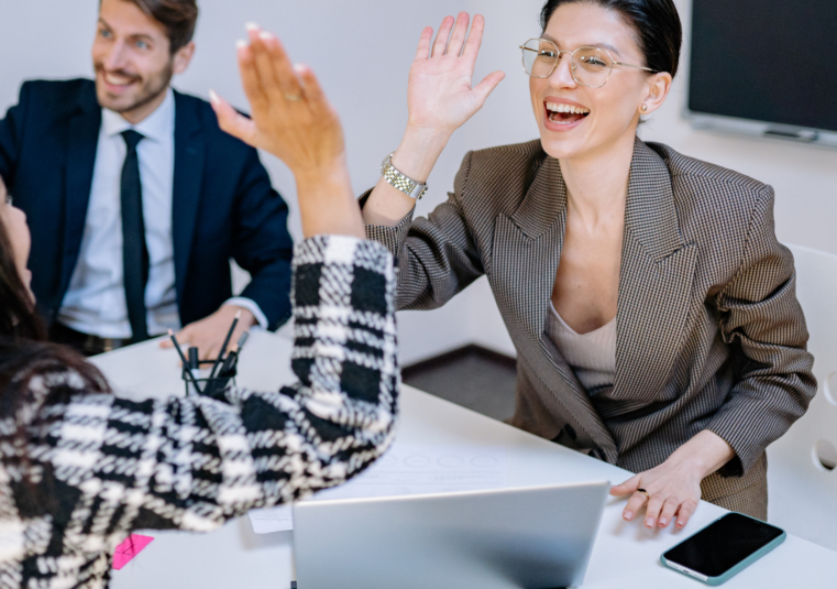 Team im Besprechungsraum eines Unternehmens feiert Fortschritt in einem Meeting, Kolleginnen klatschen sich ab während ein Kollege im Anzug lächelt